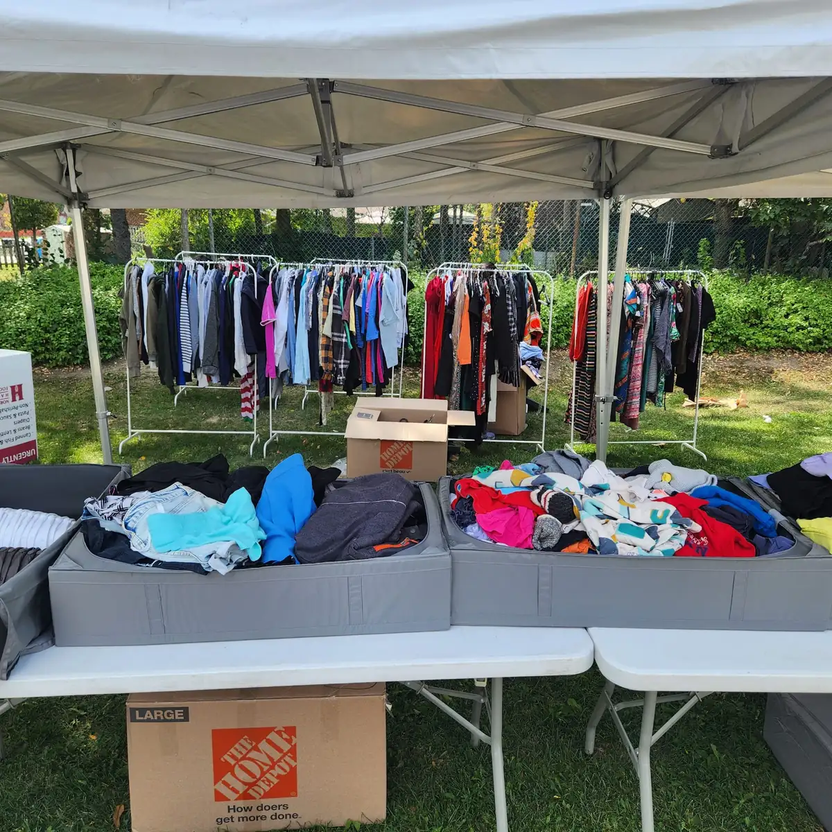 Wide shot of a white canopy tent with clothing racks, folding tables and donation boxes at an outdoor drive